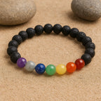 7 Chakra bracelet made with colorful gemstone beads and black lava stones placed on sand background.